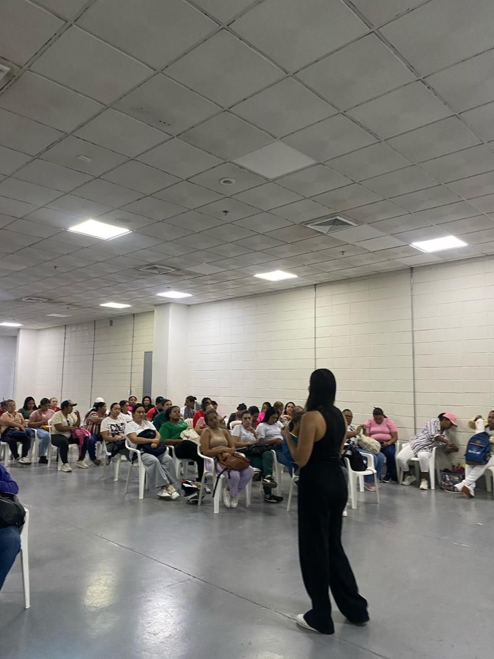 Una facilitadora de pie habla a un grupo numeroso de mujeres sentadas en sillas plásticas en un salón amplio.