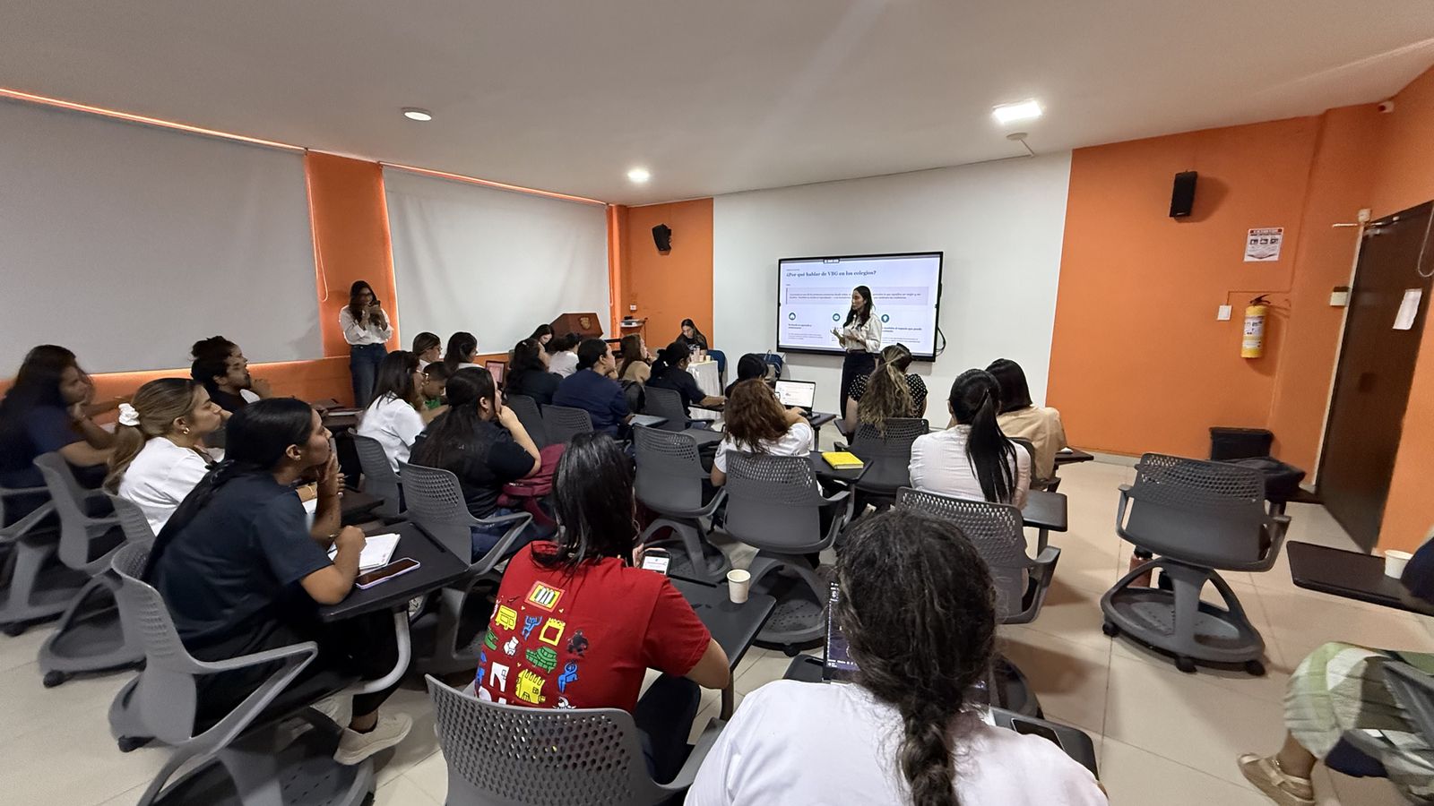 Vista amplia de un salón con participantes sentados atendiendo a una facilitadora junto a la pantalla.