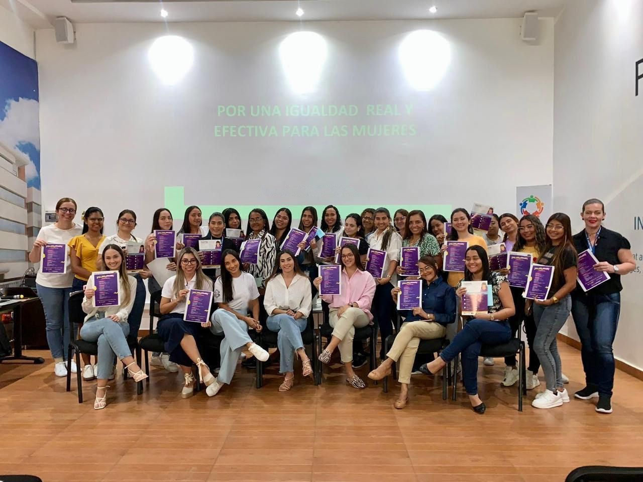 Grupo de unas treinta mujeres sonrientes muestran folletos morados frente a una pantalla que dice «Por una igualdad real y efectiva para las mujeres».