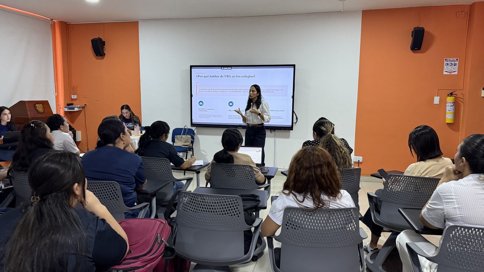 Una facilitadora explica una presentación frente a un salón de clase lleno de participantes.