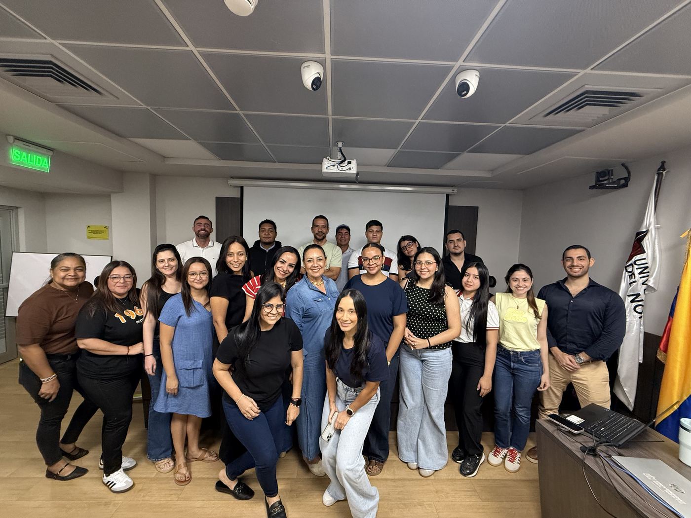 Grupo de participantes sonrientes posan en un aula junto a la bandera de la Universidad del Norte.